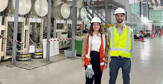 Female and male cadets wearing safety vests and hard hats stand in factory